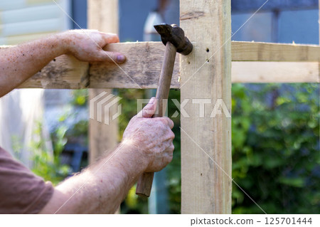 Close-up of hands of man with vitiligo using hammer to assemble wooden framework, showing focused effort in woodworking or construction outdoors Close-up of hands of man with vitiligo using hammer to assemble wooden framework, showing focused effort in woodworking or construction outdoors 125701444