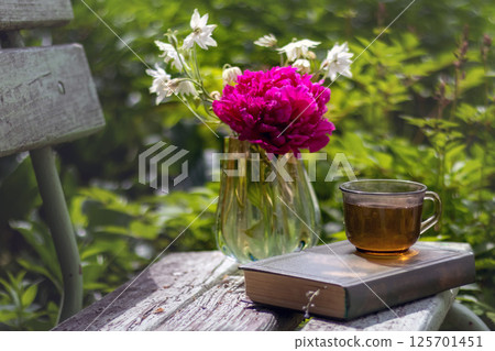 Quiet garden setting with glass of tea, book, and bright bouquet of purple peonies in vase on rustic wooden bench surrounded by lush greenery 125701451