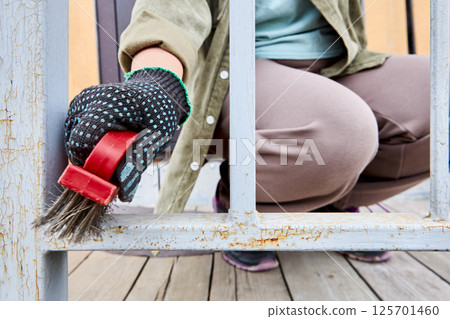 Wire brush is being used by person wearing dotted glove to remove rust from metal railing during surface preparation process on wooden deck. 125701460