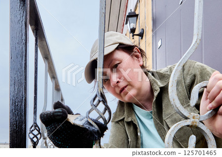 Mature woman carefully applies black paint to detailed metal railings of front porch using precise brush technique. 125701465
