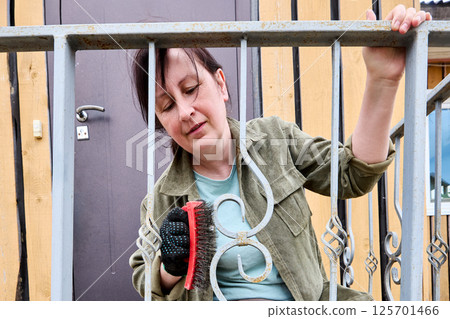 Wire brush is held by middle aged woman cleaning decorative scrollwork on metal railing during surface preparation on porch outside wooden house. 125701466