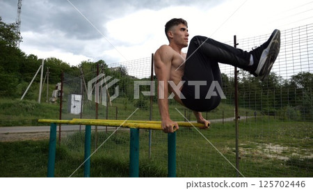 Strong guy training abdominal muscles on parallel bars at sports ground. Young shirtless sportsman exercising at nature. Sporty athlete working out at nature. Concept of active sportive lifestyle Strong guy training abdominal muscles on parallel bars at sports ground. Young shirtless sportsman exercising at nature. Sporty athlete working out at nature. Concept of active sportive lifestyle 125702446