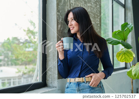 Portrait of beautiful relaxed brunette 30s woman with cup in hands looking out window Portrait of beautiful relaxed brunette 30s woman with cup in hands looking out window 125702537
