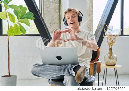 Happy teenage boy with headphones using laptop for video chat, making heart gesture 125702621