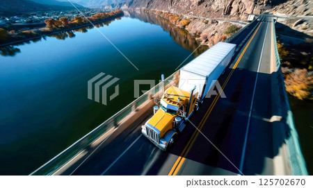 A bright yellow semi-truck driving across a scenic bridge over a calm river with mountains and autumn trees in the background 125702670