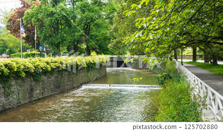 Old waterfall in the summer city with old bridge. Nature background. Falls with clean water in sunny day and green grass around. Old waterfall in the summer city with old bridge. Nature background. Falls with clean water in sunny day and green grass around. 125702880
