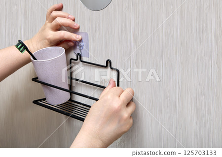 Hands are installing black wire bathroom shelf on wall using transparent nano adhesive hook for easy and damage-free bathroom hardware fitting. 125703133