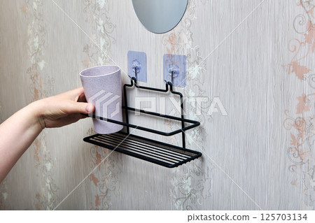 Hand places plastic cup onto black wire bathroom shelf fixed to wall with two transparent nano adhesive hooks for convenient storage solution. Hand places plastic cup onto black wire bathroom shelf fixed to wall with two transparent nano adhesive hooks for convenient storage solution. 125703134