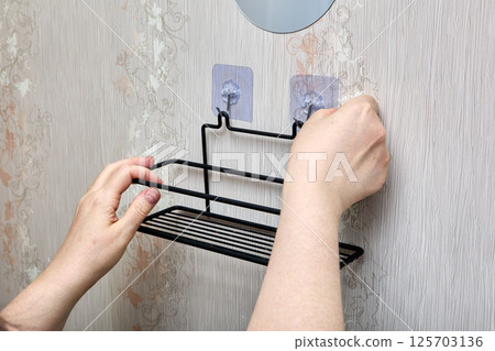Black wire bathroom rack is being installed on patterned wall using two transparent adhesive hooks for secure and damage-free fitting. Black wire bathroom rack is being installed on patterned wall using two transparent adhesive hooks for secure and damage-free fitting. 125703136