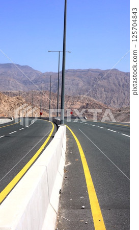 Road snakes beside bare ridges, asphalt smooth against desert stone contrast, distant peaks faded in haze, wild bushes sparse on slope. Mountain desert passage. 125704843