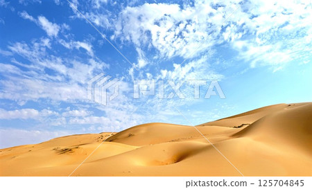 Golden sand dunes undulating across desert landscape, casting intricate shadows beneath expansive azure sky with delicate white clouds, revealing majestic arid terrain's serene beauty Golden sand dunes undulating across desert landscape, casting intricate shadows beneath expansive azure sky with delicate white clouds, revealing majestic arid terrain's serene beauty 125704845