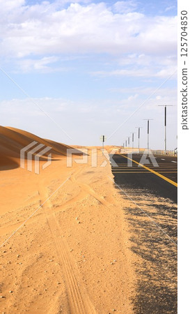 Tire marks fade into golden dunes beside paved road, quiet trace of motion carved in sand speaks of passage, solitude, and shifting time, iconic desert tourist attraction. 125704850