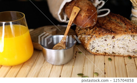 Freshly baked seeded bread in burlap sack, metal bowl with salt, wooden spoon, glass of orange juice on rustic wooden surface, creating wholesome, natural breakfast scene. Freshly baked seeded bread in burlap sack, metal bowl with salt, wooden spoon, glass of orange juice on rustic wooden surface, creating wholesome, natural breakfast scene. 125704858