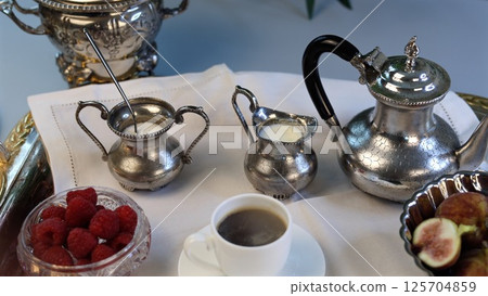 Breakfast arranged with intricate silverware, black-handled teapot, crystal bowl filled with vibrant raspberries, figs sliced into elegant portions, and smooth white porcelain coffee cup. Breakfast arranged with intricate silverware, black-handled teapot, crystal bowl filled with vibrant raspberries, figs sliced into elegant portions, and smooth white porcelain coffee cup. 125704859