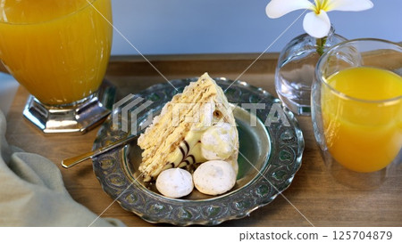 Sweet breakfast, minimalist design with silver plate reflecting pale yellow cake, vibrant orange juice in glass, white meringues, blending simplicity with striking colors. Sweet breakfast, minimalist design with silver plate reflecting pale yellow cake, vibrant orange juice in glass, white meringues, blending simplicity with striking colors. 125704879