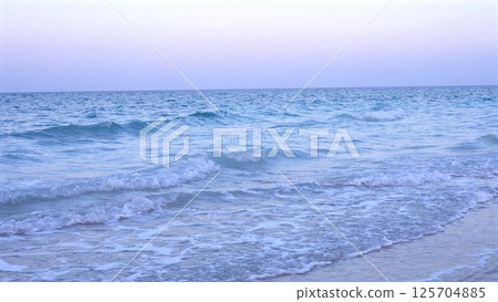 Soft blue ocean waves edged in white foam glide gently onto golden sand as sky begins to lighten, capturing serene stillness of early hours before sunrise. Early peace concept. 125704885
