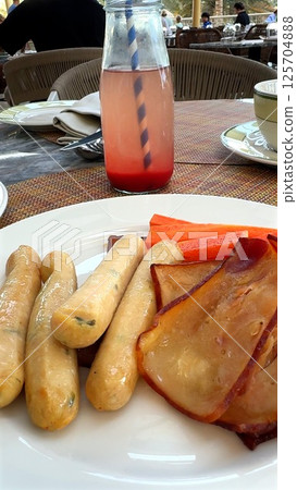 Sausages arranged beside smoked meat slices and red drink in glass bottle create flavorful breakfast setting, bright striped straw and wicker seating complete sunny outdoor . Hearty brunch. 125704888