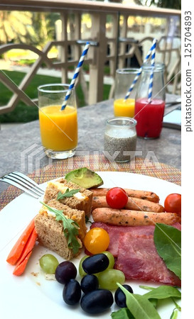 Gourmet breakfast on balcony at prestigious tourist destination, sausages and sandwiches artfully arranged, fresh juice highlights leisurely luxury vacation, ideal travel lifestyle capture. Keywords: 125704893