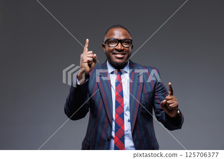 Confident businessman in striped suit and glasses, smiling and pointing upward with both hands. Concept of idea, success, motivation, leadership, and inspiration. Studio shot, gray background. 125706377
