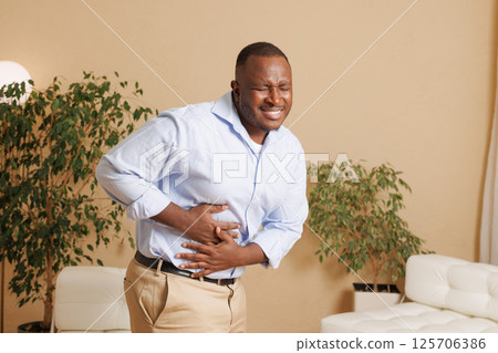 Man suffering from severe stomach pain at home. He clutches his abdomen with a pained expression, indicating digestive issues, cramps, or food poisoning. Health and medical concept. 125706386