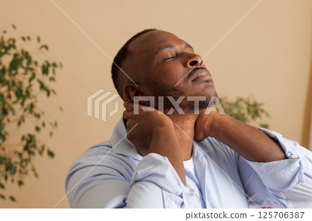 Close-up of a man experiencing neck pain, holding the back of his neck with both hands, eyes closed in discomfort. Concept of muscle strain, tension, or cervical spine issues. Health problem. 125706387