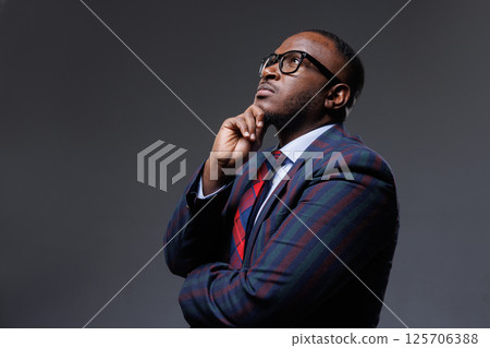 Thoughtful businessman in striped suit and glasses, looking up with hand on chin. Concept of decision-making, planning, strategy, leadership, and innovation. Dark studio background. 125706388