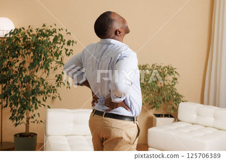 Man experiencing severe lower back pain at home. He holds his back with both hands, grimacing in discomfort. Concept of backache, muscle strain, or injury. Health and medical issue. 125706389
