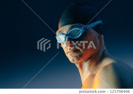 Focused male swimmer in swim cap and goggles, wet skin glowing under dramatic lighting, ready to compete. 125706406