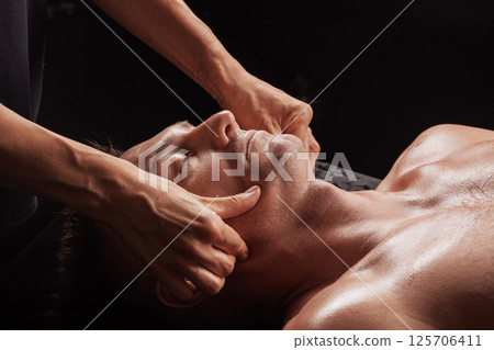 Close-up of facial massage technique with thumbs on jawline of relaxed man during spa therapy session. Close-up of facial massage technique with thumbs on jawline of relaxed man during spa therapy session. 125706411
