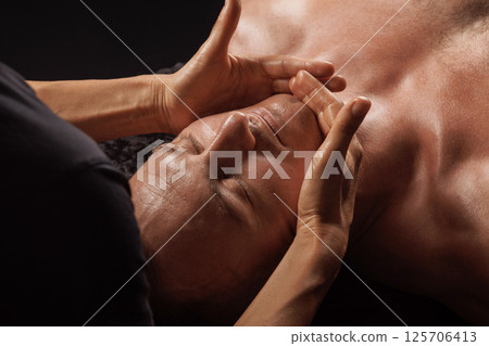 Therapist performing facial massage on a relaxed man in spa setting, focusing on jaw and chin muscle areas. Therapist performing facial massage on a relaxed man in spa setting, focusing on jaw and chin muscle areas. 125706413