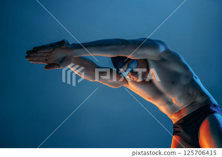 Professional male swimmer in dive pose, arms extended, under blue and orange light, ready to leap into water. 125706415