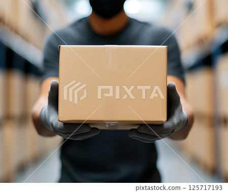 warehouse worker in protective gloves holds cardboard box, showcasing safety and diligence in storage environment. background features shelves filled with packages 125717193