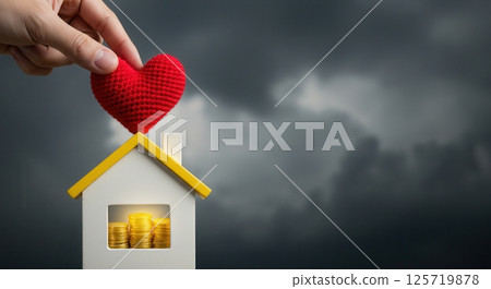 Hand places a red heart on a house model with gold coins inside, set against a dark, stormy sky. Concept for home, love, security, and financial safety. 125719878