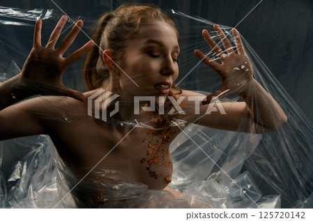 Red naked woman with golden body art poses inside transparent film Red naked woman with golden body art poses inside transparent film 125720142