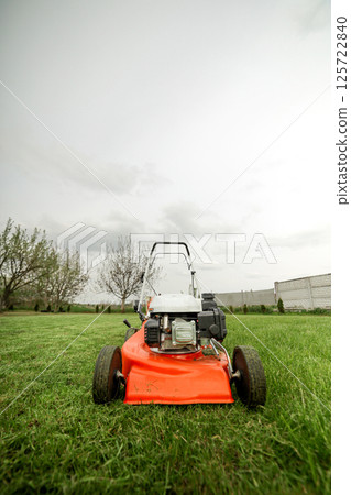 Gardening work tools. Wide angle view of orange electric lawn mower, wheels, motor on bright lush green grass at backyard. Rotary lawn mower machine cut lawn. Professional lawn care service. Gardening work tools. Wide angle view of orange electric lawn mower, wheels, motor on bright lush green grass at backyard. Rotary lawn mower machine cut lawn. Professional lawn care service. 125722840