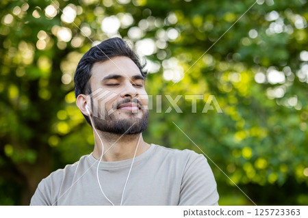 Young man with eyes closed enjoying music through headphones in outdoor park environment. Expression shows relaxation, contentment, and connection with nature. 125723363