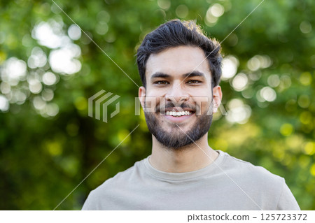 Smiling man with beard enjoying outdoors in park. Casual and relaxed expression conveys happiness and positivity. Sunlight filtering through leaves adds natural ambiance to portrait. 125723372