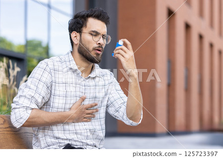 Young man seated outdoors using asthma inhaler for respiratory relief. Wearing glasses and plaid shirt, holding inhaler and chest. Outdoor urban environment with greenery and buildings in background. Young man seated outdoors using asthma inhaler for respiratory relief. Wearing glasses and plaid shirt, holding inhaler and chest. Outdoor urban environment with greenery and buildings in background. 125723397