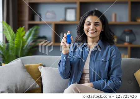 Portrait of a young smiling Muslim woman sitting on the sofa at home in a shirt, holding an inhaler for breathing, and showing a bottle of medicine to the camera. Portrait of a young smiling Muslim woman sitting on the sofa at home in a shirt, holding an inhaler for breathing, and showing a bottle of medicine to the camera. 125723420