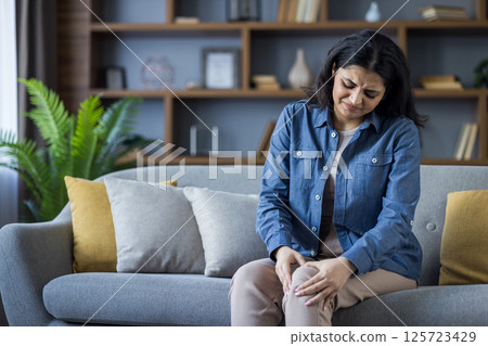 A young Muslim woman sits on the couch at home and holds her leg with her hands, feeling severe pain in her knee and bones. A young Muslim woman sits on the couch at home and holds her leg with her hands, feeling severe pain in her knee and bones. 125723429