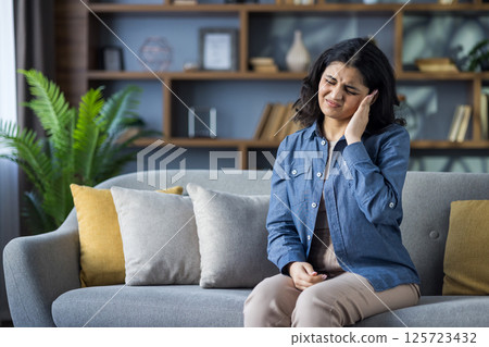 Muslim young woman in shirt sitting on sofa at home, grimacing in pain, holding hand to head, suffering from pressure. Muslim young woman in shirt sitting on sofa at home, grimacing in pain, holding hand to head, suffering from pressure. 125723432