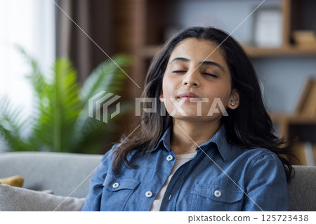 Young woman relaxing at home on the couch, eyes closed, practicing meditation and mindfulness for stress relief in a cozy living room. Young woman relaxing at home on the couch, eyes closed, practicing meditation and mindfulness for stress relief in a cozy living room. 125723438