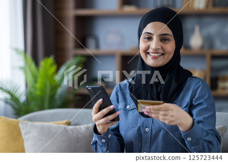 Portrait of a young Islamic woman in a hijab sitting on the sofa at home, holding a credit card and a mobile phone, looking at the camera with a smile. 125723444