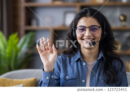 Friendly young woman wearing a headset smiles and waves during a virtual call, creating a warm and professional connection from home. Friendly young woman wearing a headset smiles and waves during a virtual call, creating a warm and professional connection from home. 125723451