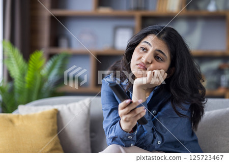 A young woman looks bored while sitting on the couch in her living room, holding a tv remote while watching television. 125723467
