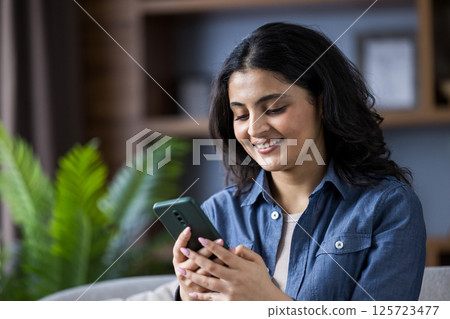 Young Indian woman in denim shirt is at home, sitting on the couch and smiling, using her mobile phone. Close-up photo. 125723477