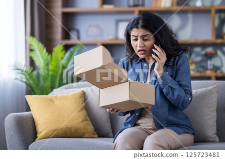 A young woman looks shocked as she opens a cardboard package, immediately calling customer service to report the issue at her home. A young woman looks shocked as she opens a cardboard package, immediately calling customer service to report the issue at her home. 125723481