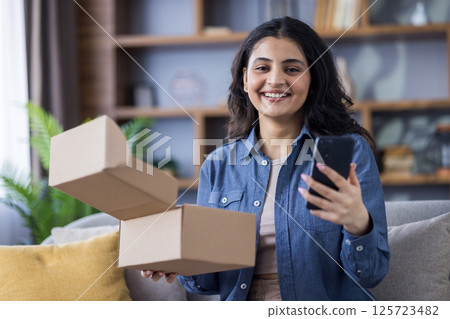 A young woman is sitting at home, excited to receive her recent order and holding her smartphone for sharing online. A young woman is sitting at home, excited to receive her recent order and holding her smartphone for sharing online. 125723482
