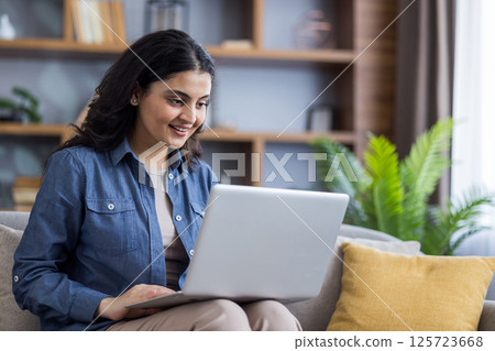 A young woman in a denim shirt smiles while using her laptop on the sofa at home, staying connected and productive. A young woman in a denim shirt smiles while using her laptop on the sofa at home, staying connected and productive. 125723668