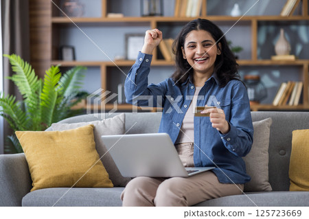 A young Indian woman celebrates an online purchase with a credit card while sitting on her sofa with her laptop in a cozy living room. 125723669